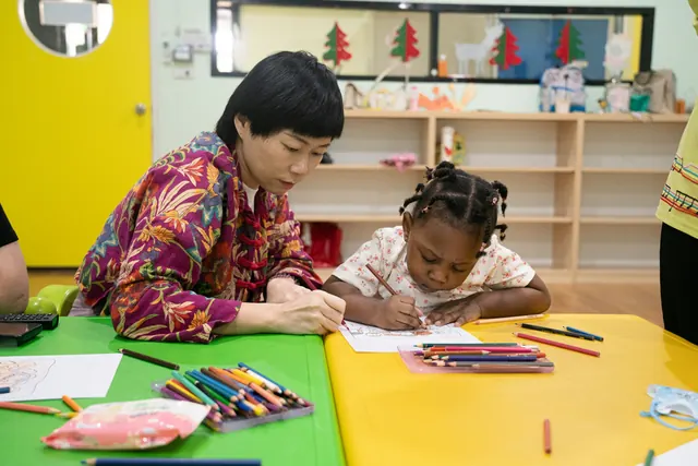 A woman and a young girl draw side by side at a bright classroom table.