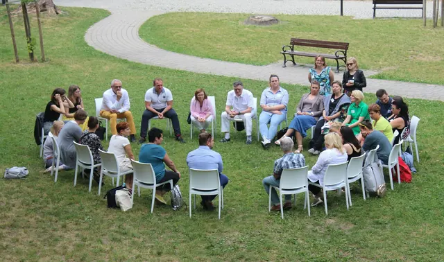 A group of people sits in a wide circle of chairs on a lawn, talking together.