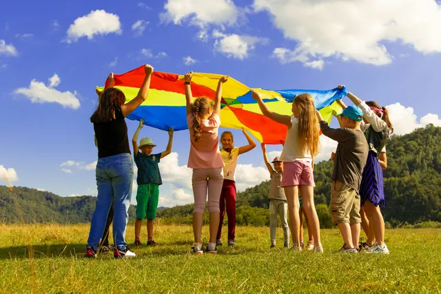 Children and adults lift a bright play parachute together in a sunny field.