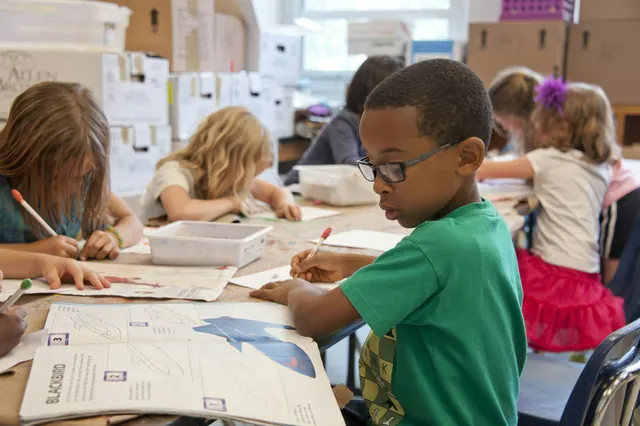 Children draw and write together at their tables in a bright primary school classroom.