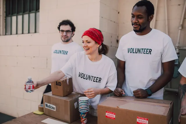 Three smiling volunteers in matching volunteer t-shirts work together at a donation table.