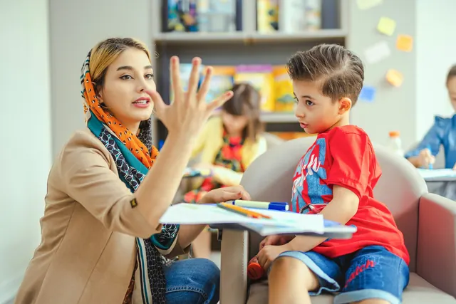 A teacher counts on her fingers with a young boy during a lesson in a bright classroom.