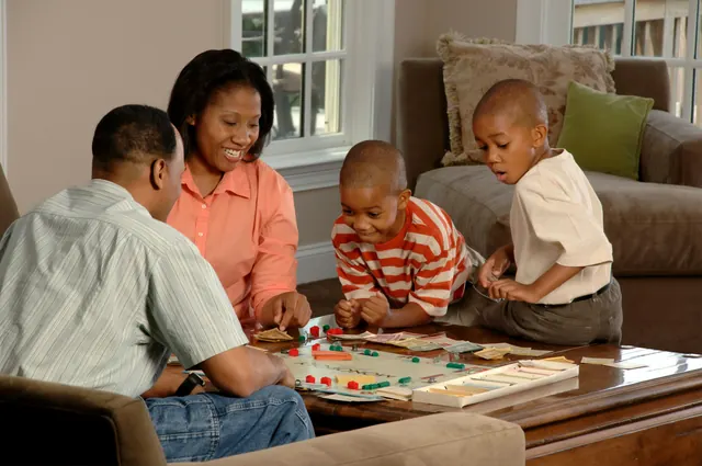 Two parents and their two young sons play a board game together around the living room table.
